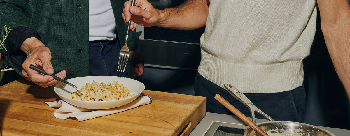 Two people's hands hold forks over a white plate of cavatappi pasta resting on a wooden cutting board with a cloth napkin, with kitchen tongs and a pan visible nearby.