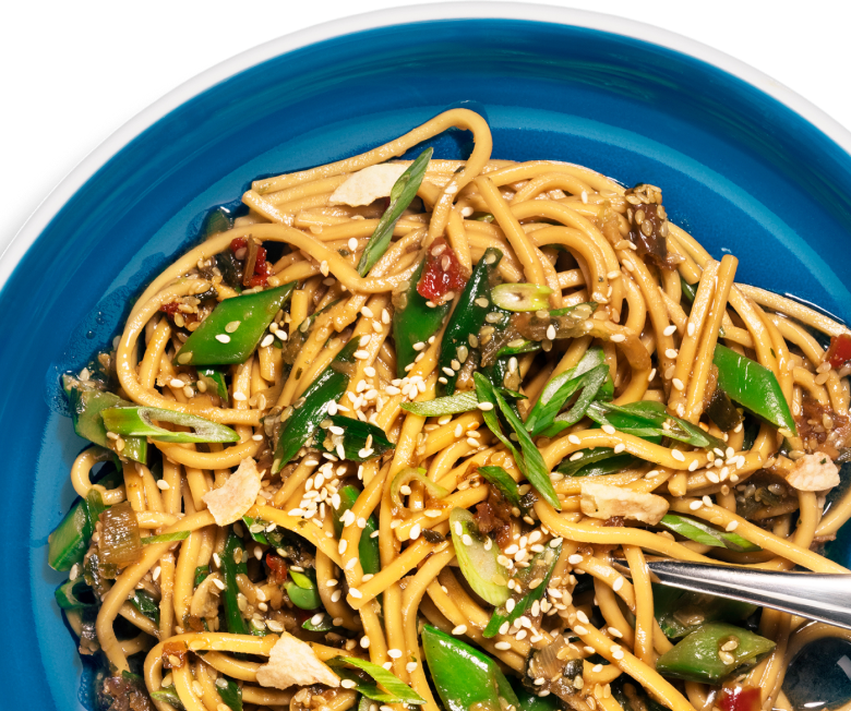 Overhead close-up of Riff Sesame Ginger Noodles topped with sesame seeds, sliced scallions, and snap peas in a blue bowl, with a fork resting on the side.