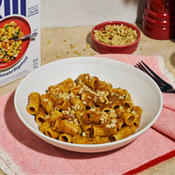 A box of Riff Spicy Tomato Rigatoni chef-crafted meal kit next to a white bowl of the plated rigatoni topped with crunchy breadcrumbs, a small red bowl of extra crunchy topping, a black fork on a red napkin, and a red pepper grinder.