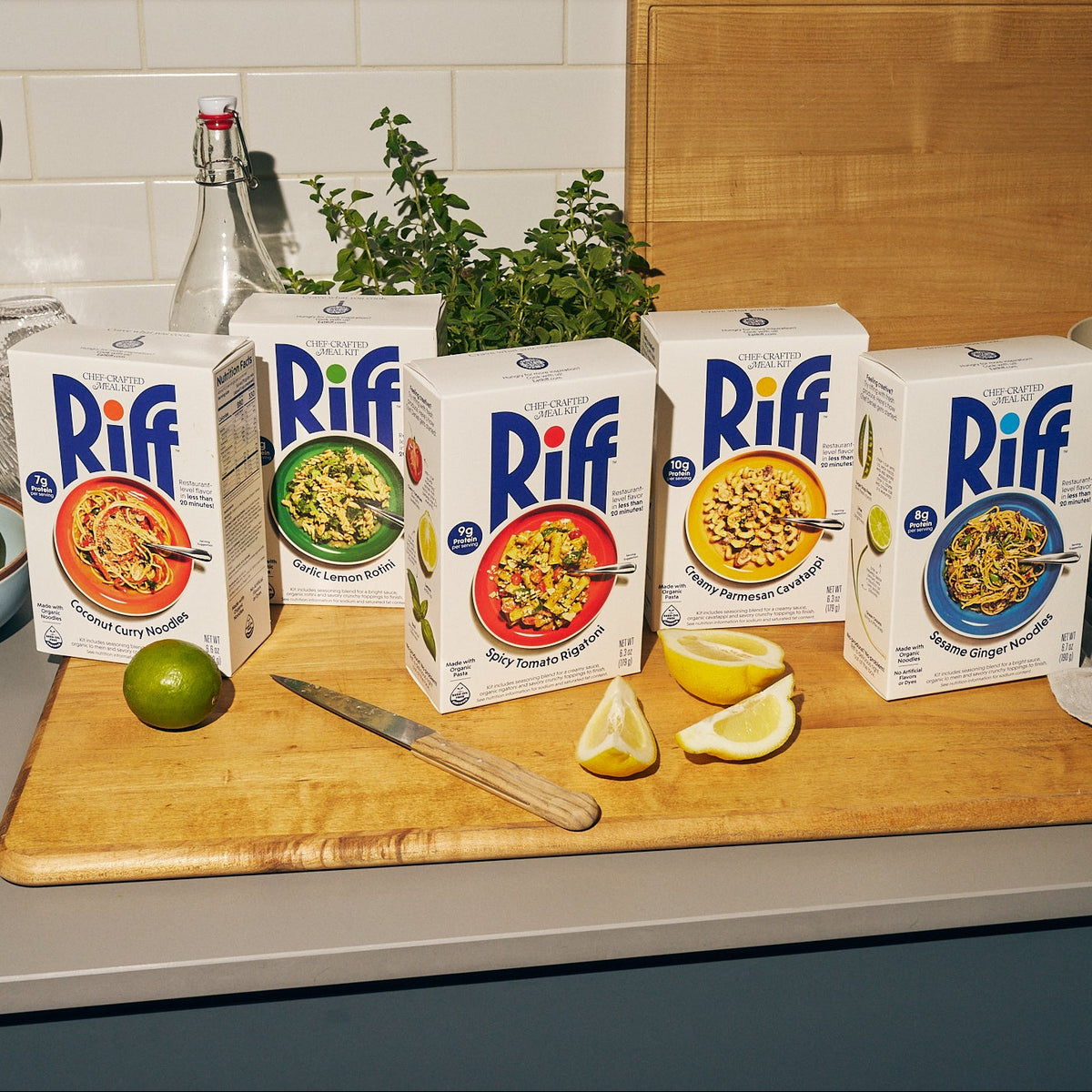 Five Riff chef-crafted meal kit boxes lined up on a wooden cutting board: Coconut Curry Noodles, Garlic Lemon Rotini, Spicy Tomato Rigatoni, Creamy Parmesan Cavatappi, and Sesame Ginger Noodles, each showing its plated dish, with a lime, a paring knife, lemon wedges, and a potted herb plant nearby.