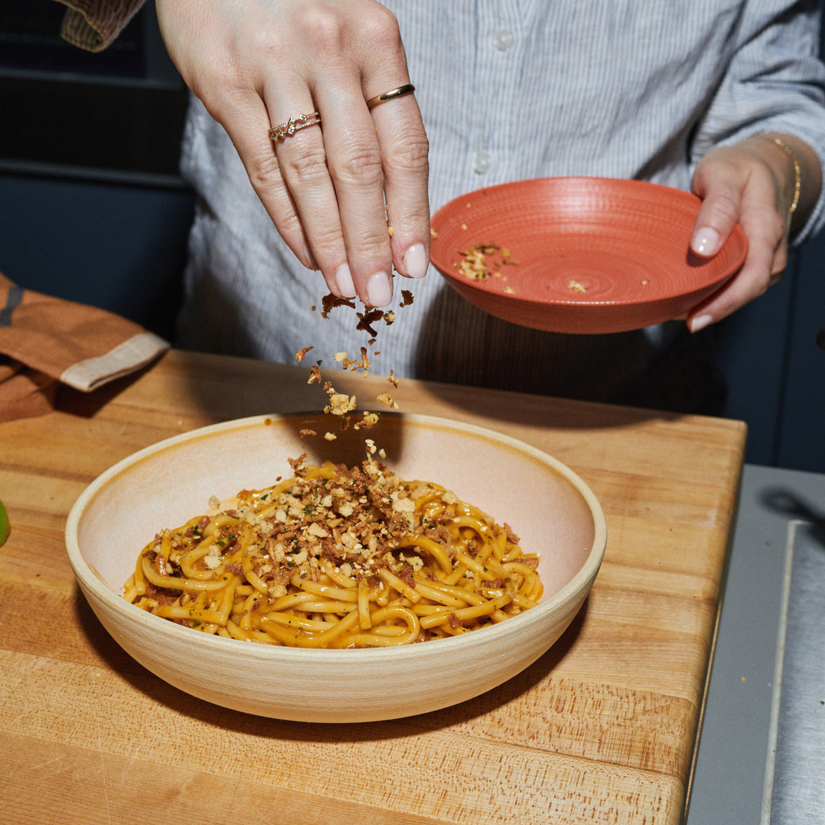 A hand sprinkles crunchy topping from a red bowl onto a bowl of Riff Coconut Curry Noodles on a wooden cutting board.