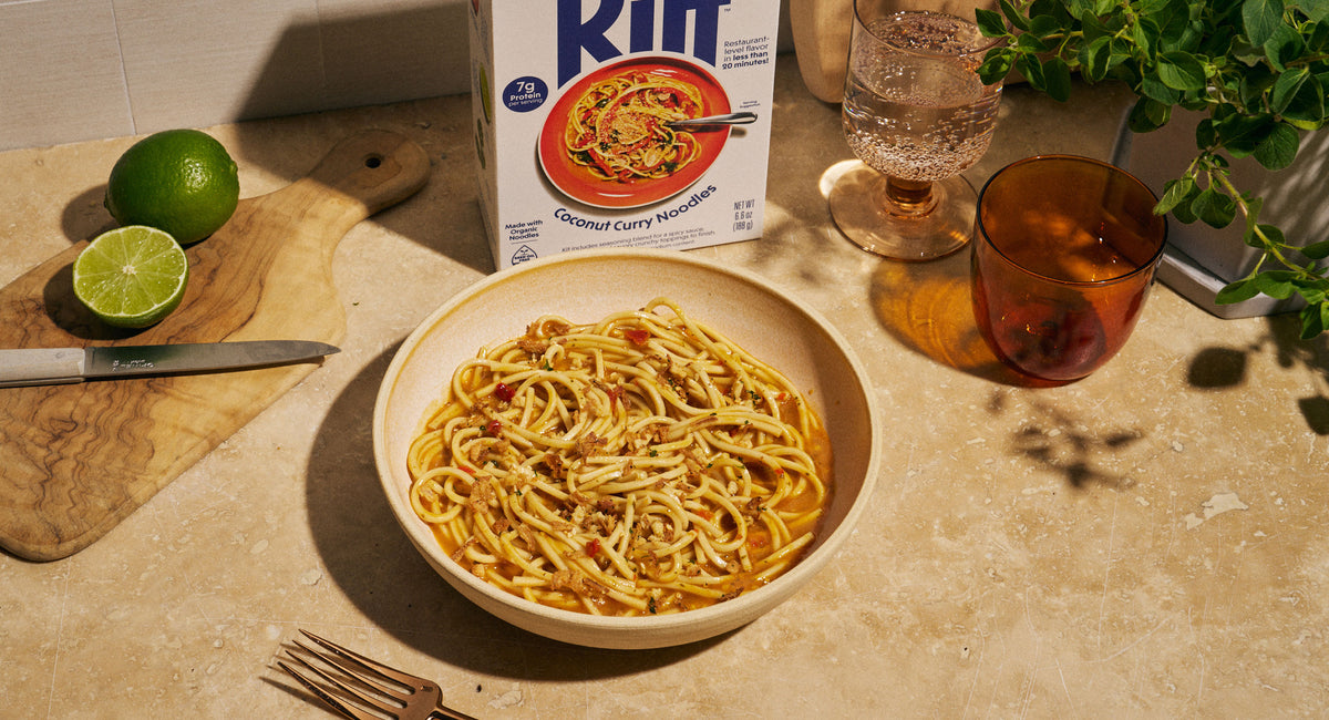 A bowl of Riff Coconut Curry Noodles sits on a kitchen counter next to the Riff box, a lime cut in half, a glass of sparkling water, and an amber glass of a beverage.