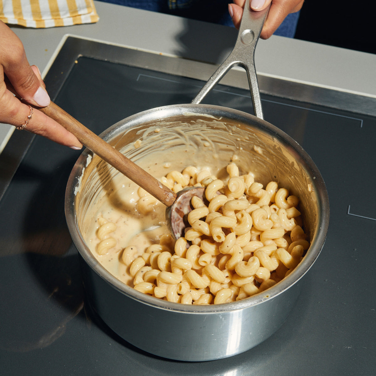 Overhead view of two hands stirring creamy cavatappi pasta in a saucepan with a wooden spoon.