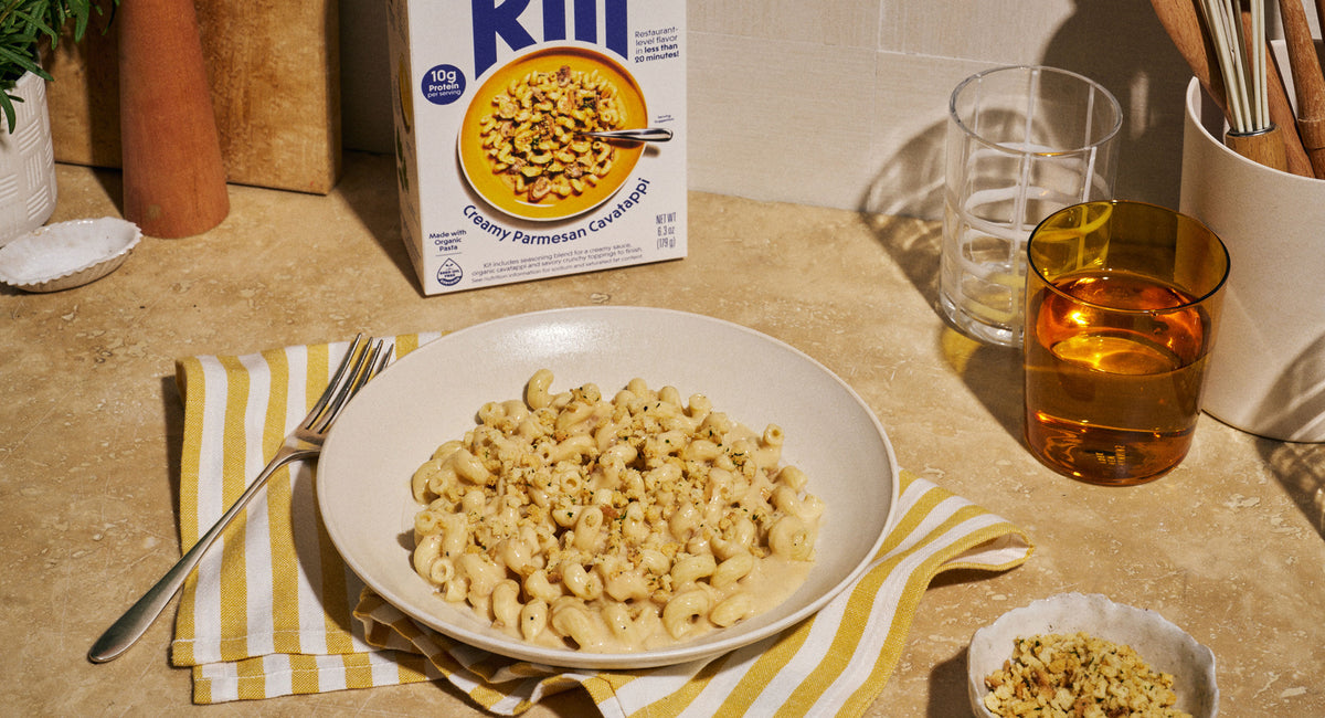 A bowl of Riff Creamy Parmesan Cavatappi topped with crunchy breadcrumbs sits on a striped napkin next to a fork, with the Riff box, glasses of water and an amber beverage, and a small dish of crunchy topping on the counter.