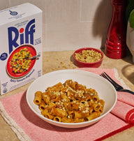A white bowl of Riff Spicy Tomato Rigatoni pasta with crumbled topping sits on a red-and-white striped napkin on a kitchen counter, next to a box of Riff Spicy Tomato Rigatoni, a small red bowl of crunchy topping, a black fork, and a red pepper mill.