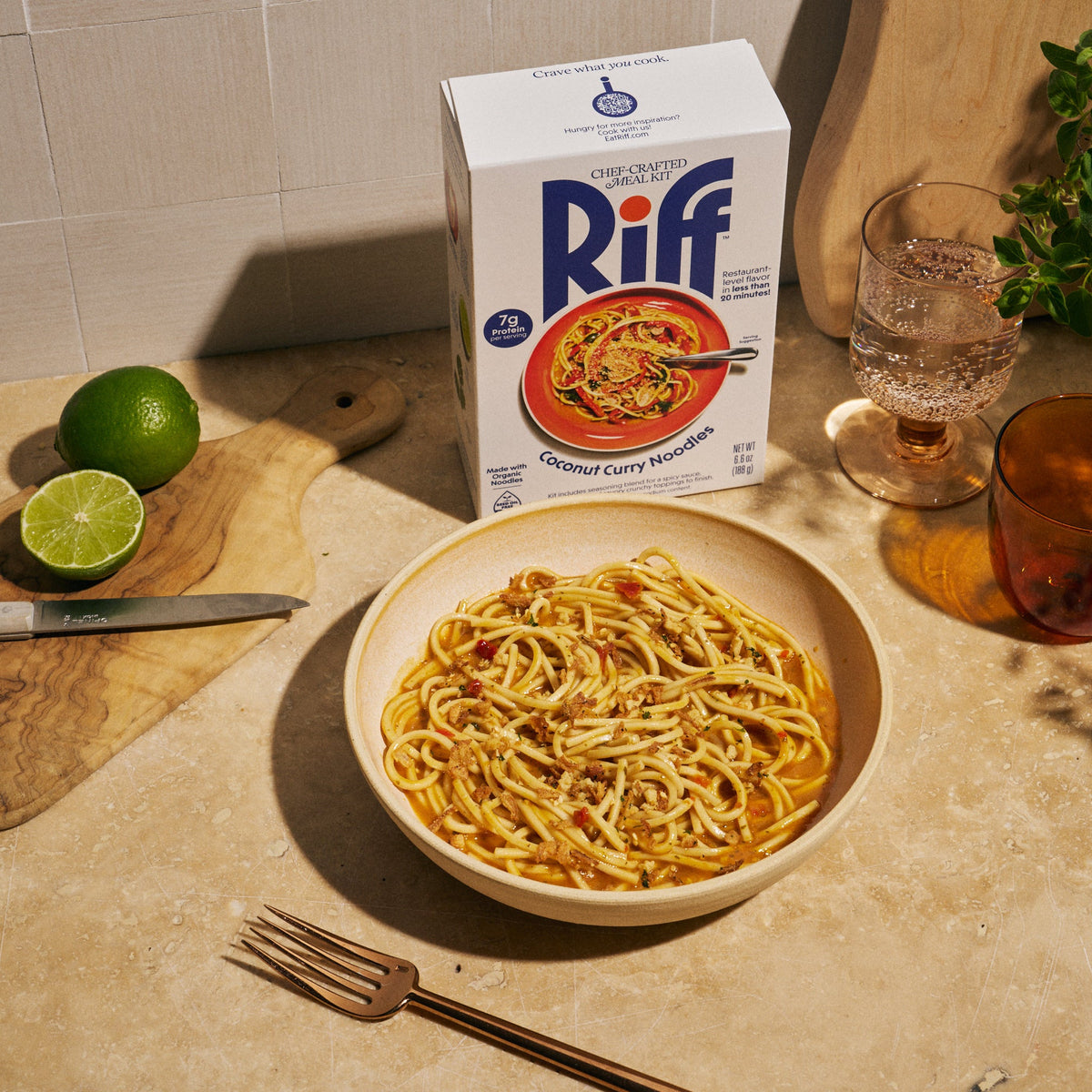 A bowl of Riff Coconut Curry Noodles sits on a kitchen counter next to the Riff box, a lime cut in half, a glass of sparkling water, and an amber glass of a beverage.