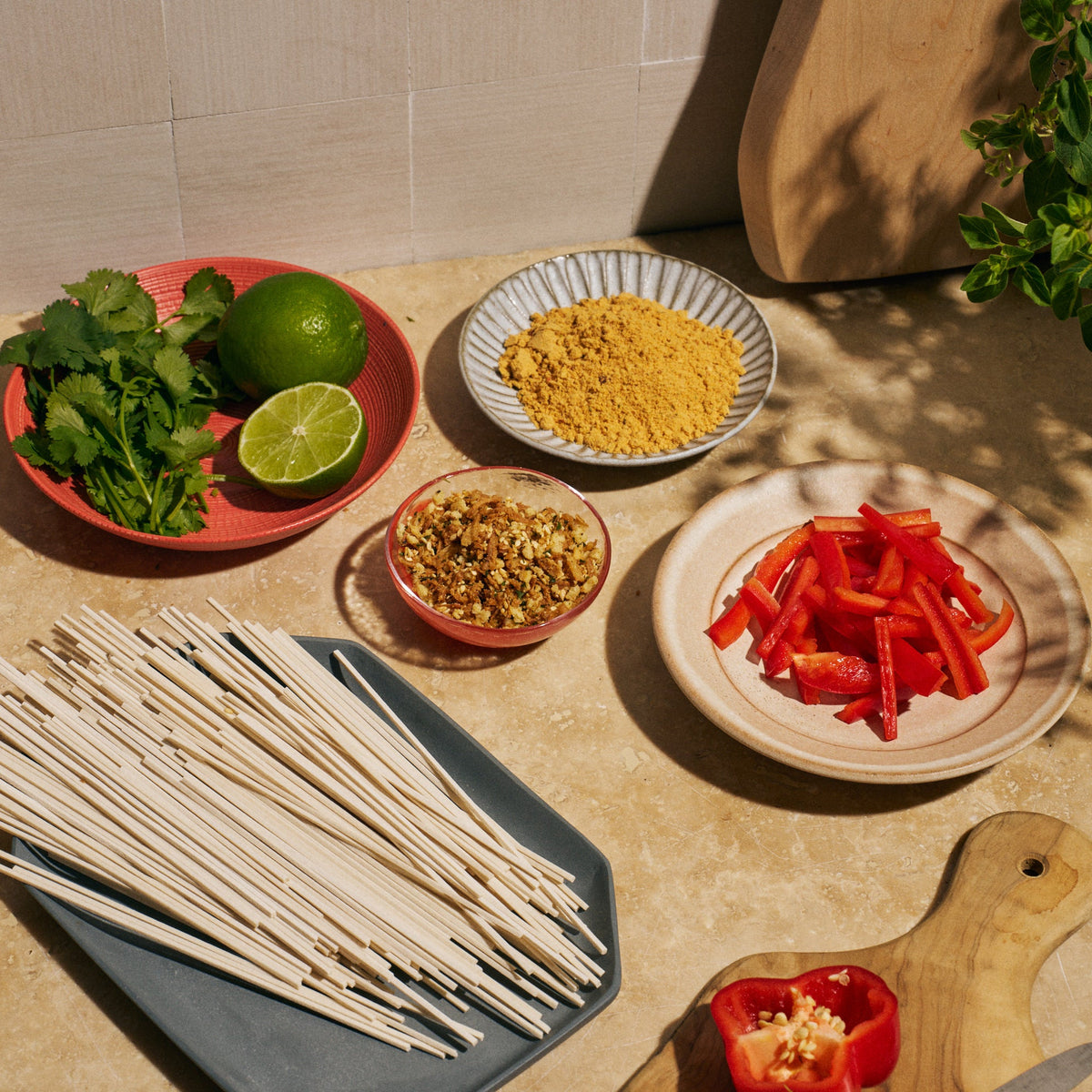 Overhead shot of ingredients on a kitchen counter for Riff Coconut Curry Noodles, including dry lo mein noodles, sliced red bell pepper, cilantro, limes, a bowl of curry seasoning, and a bowl of crunchy topping.