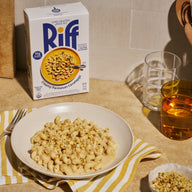A plate of Riff Creamy Parmesan Cavatappi pasta topped with crunchy breadcrumbs, set on a yellow and white striped napkin next to a box of Riff Creamy Parmesan Cavatappi chef-crafted meal kit, a fork, and glasses of water and iced tea.