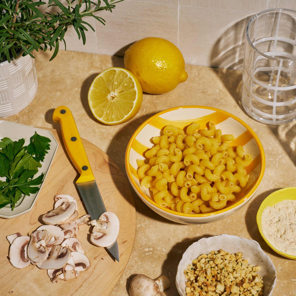 Overhead shot of raw cavatappi pasta in a yellow-striped bowl on a kitchen counter, surrounded by fresh lemons, sliced mushrooms, chopped parsley, a bowl of flour, a bowl of crunchy topping, a knife, and a small potted rosemary plant.