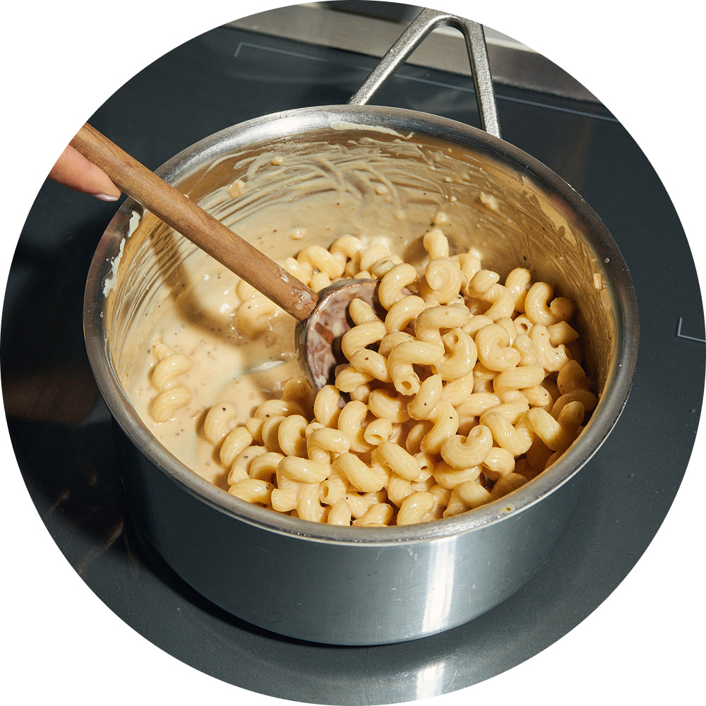 Circular overhead photo of a hand stirring cooked cavatappi pasta into a creamy sauce with a wooden spoon in a stainless steel pot.