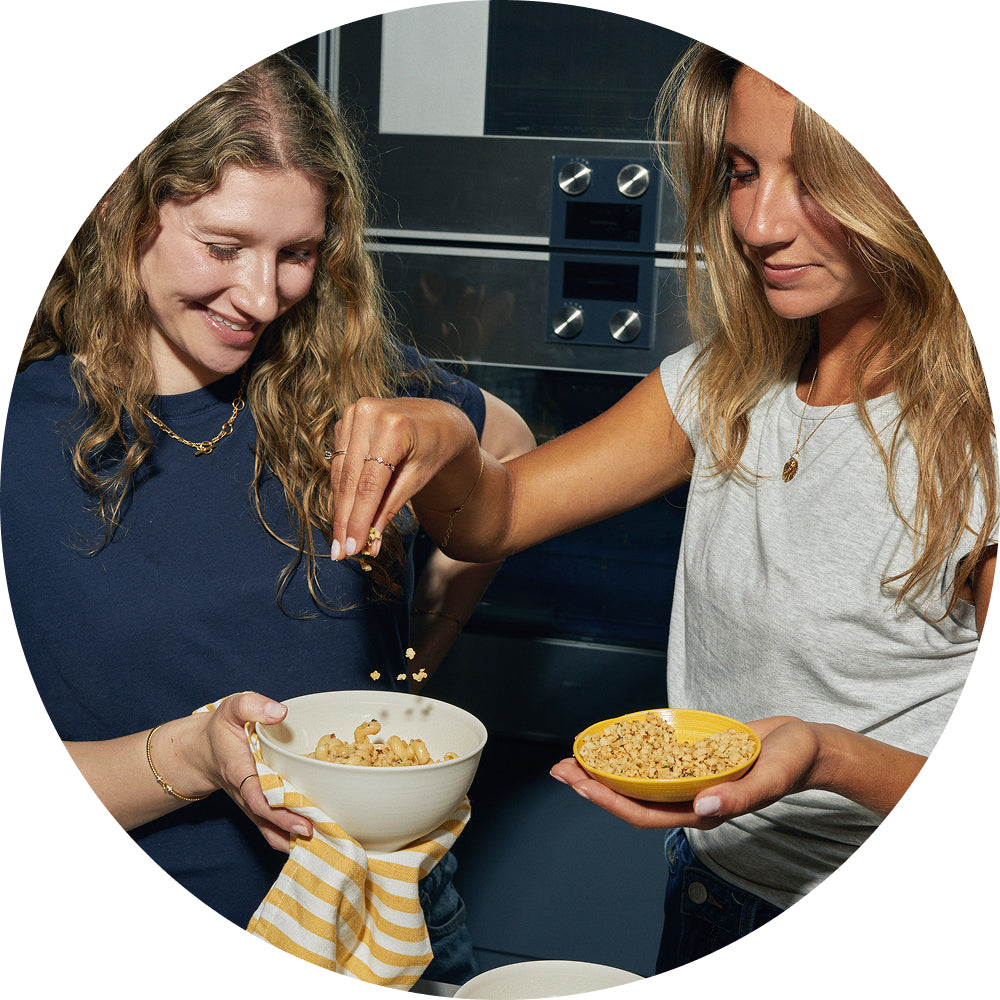 Circular photo of two women in a kitchen preparing pasta together, one sprinkling crunchy topping from her fingers into a white bowl of macaroni pasta held by the other, who also holds a yellow bowl of crunchy topping.