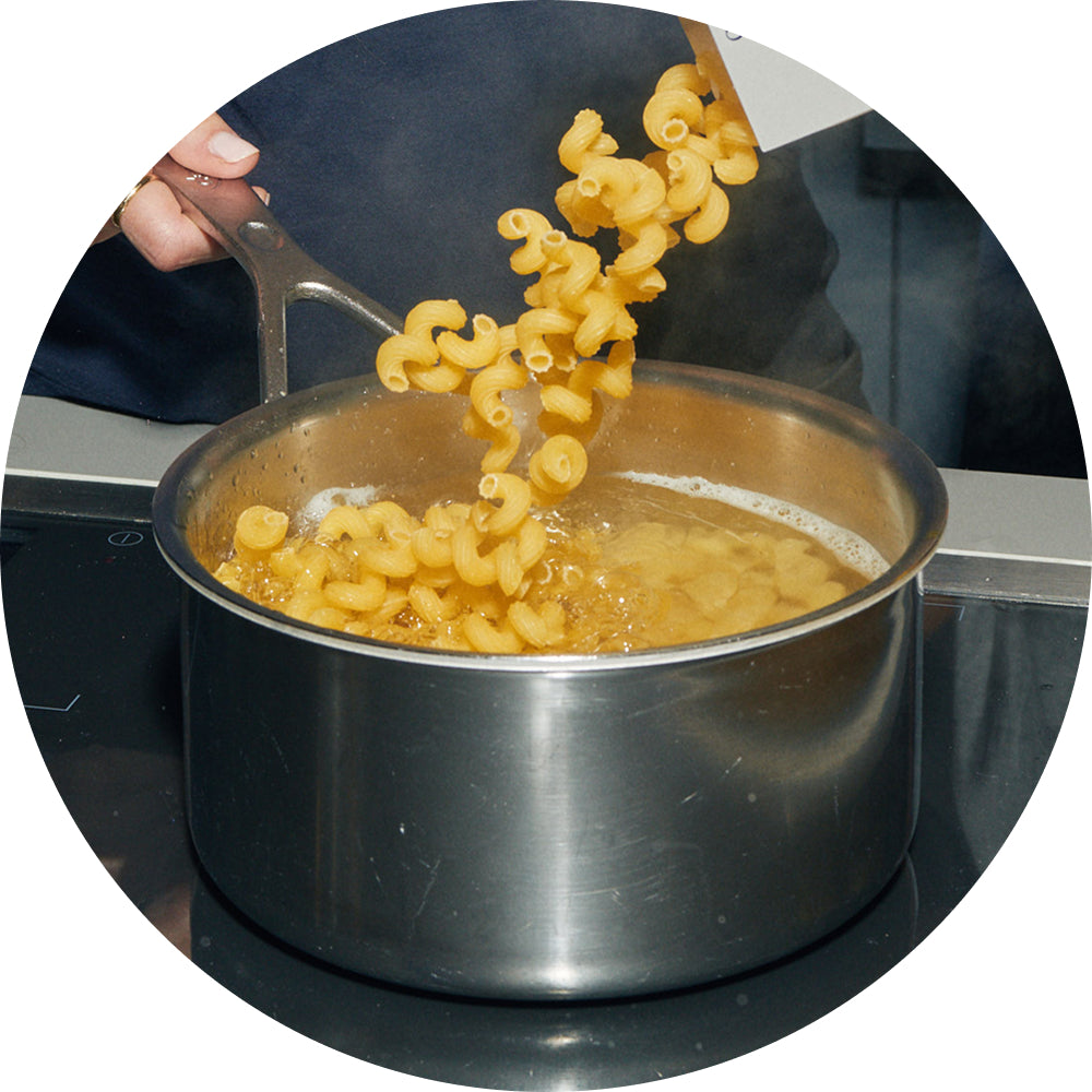 Circular photo of a hand lifting a strainer full of cooked cavatappi pasta out of a pot of boiling water on a stovetop.