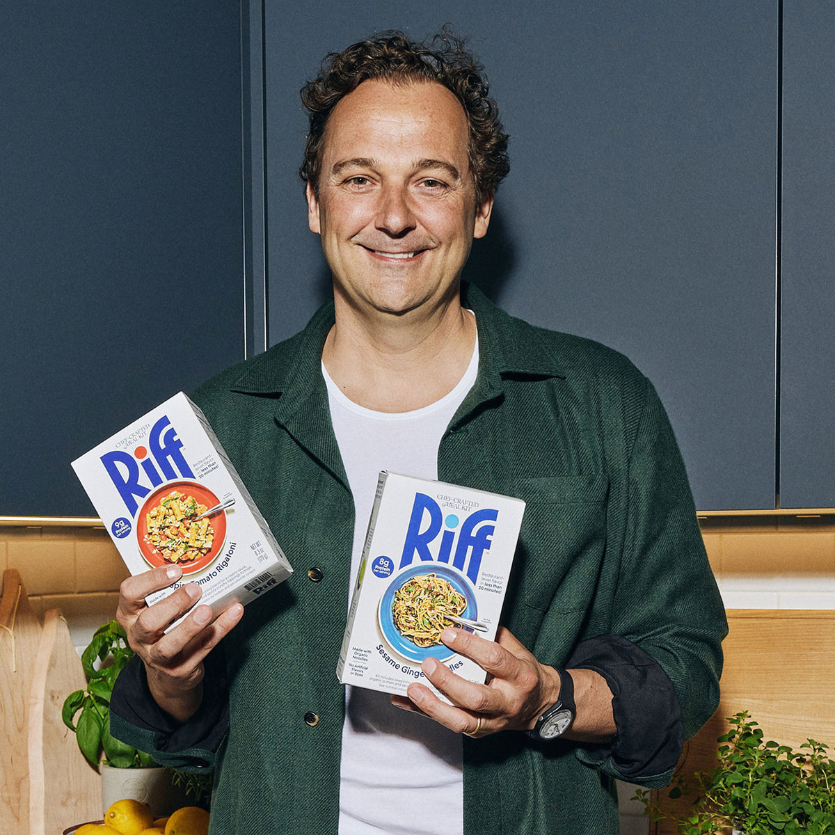A smiling man in a green jacket holds up two Riff chef-crafted meal kit boxes, Spicy Tomato Rigatoni and Sesame Ginger Noodles, in a kitchen with basil and lemons visible on the counter.