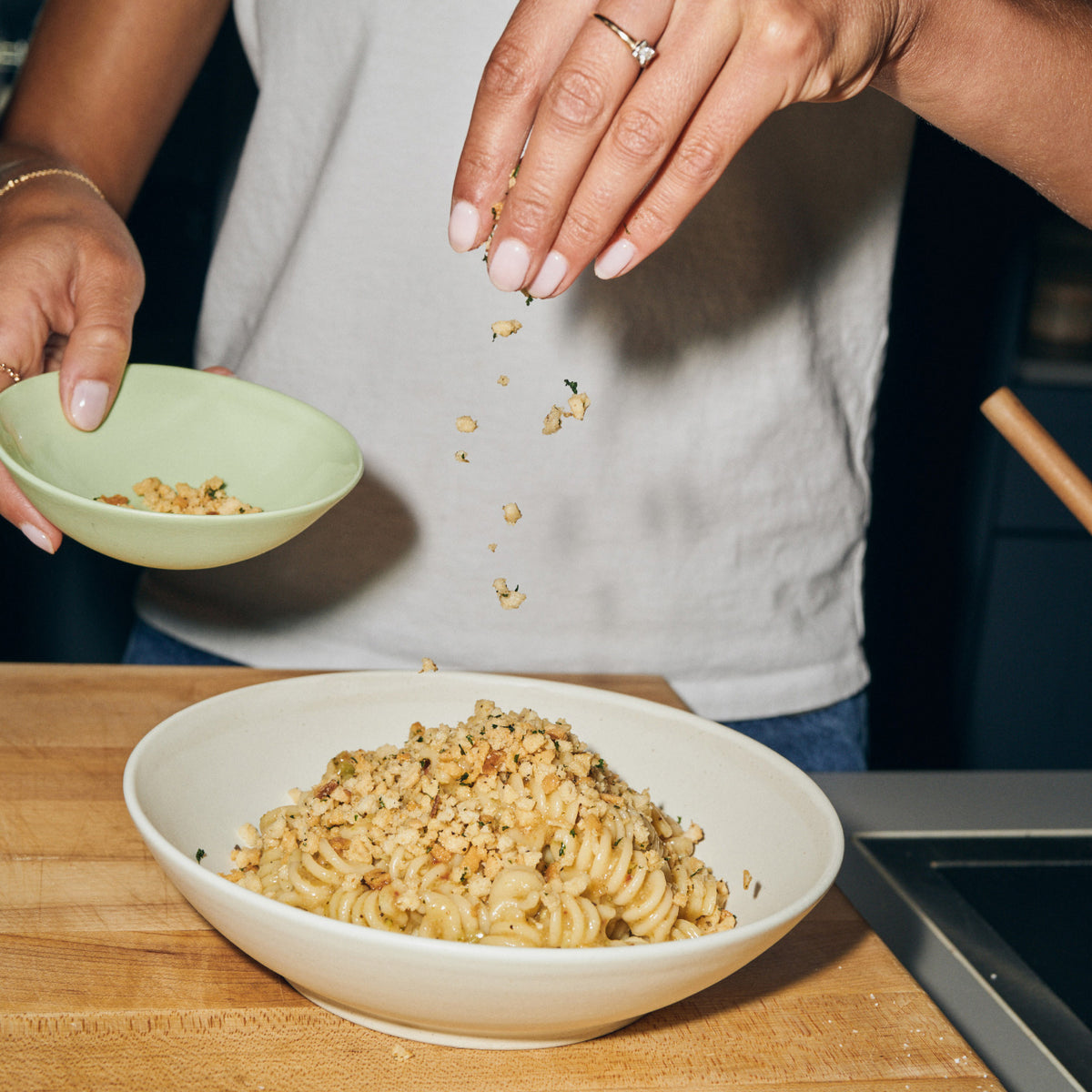 A hand sprinkles crunchy topping from a small green bowl onto a bowl of Riff Garlic Lemon Rotini on a wooden cutting board.