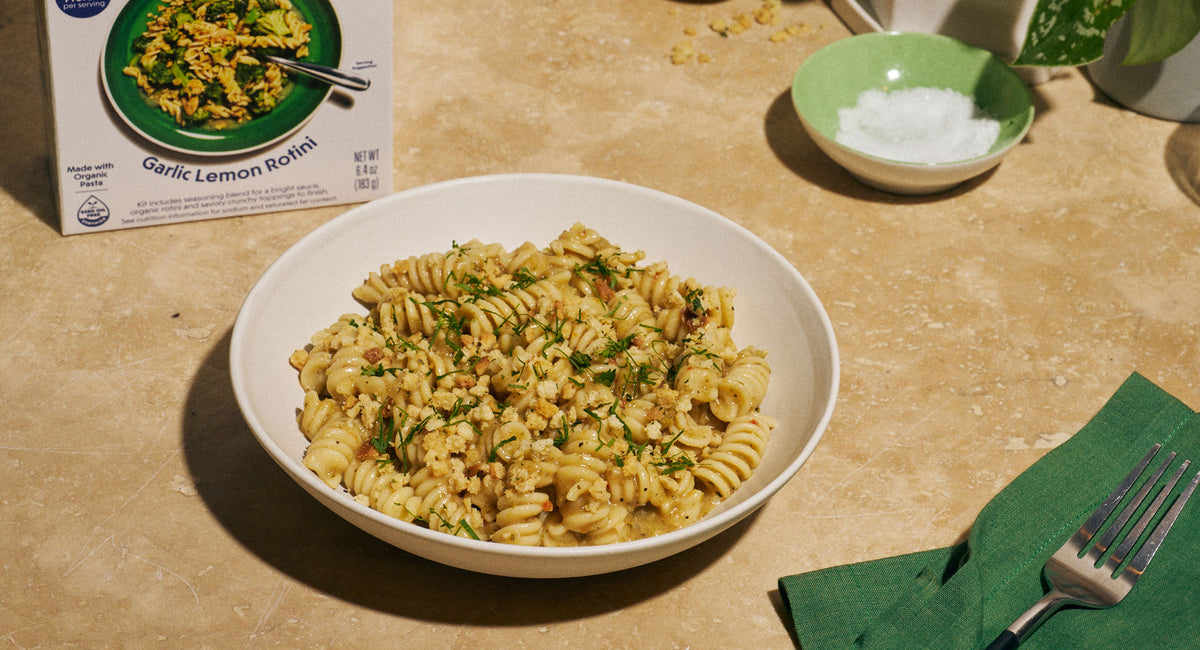 A bowl of Riff Garlic Lemon Rotini topped with crunchy breadcrumbs and fresh herbs sits on a counter next to the Riff box and a green bowl of salt, with a fork on a green napkin.