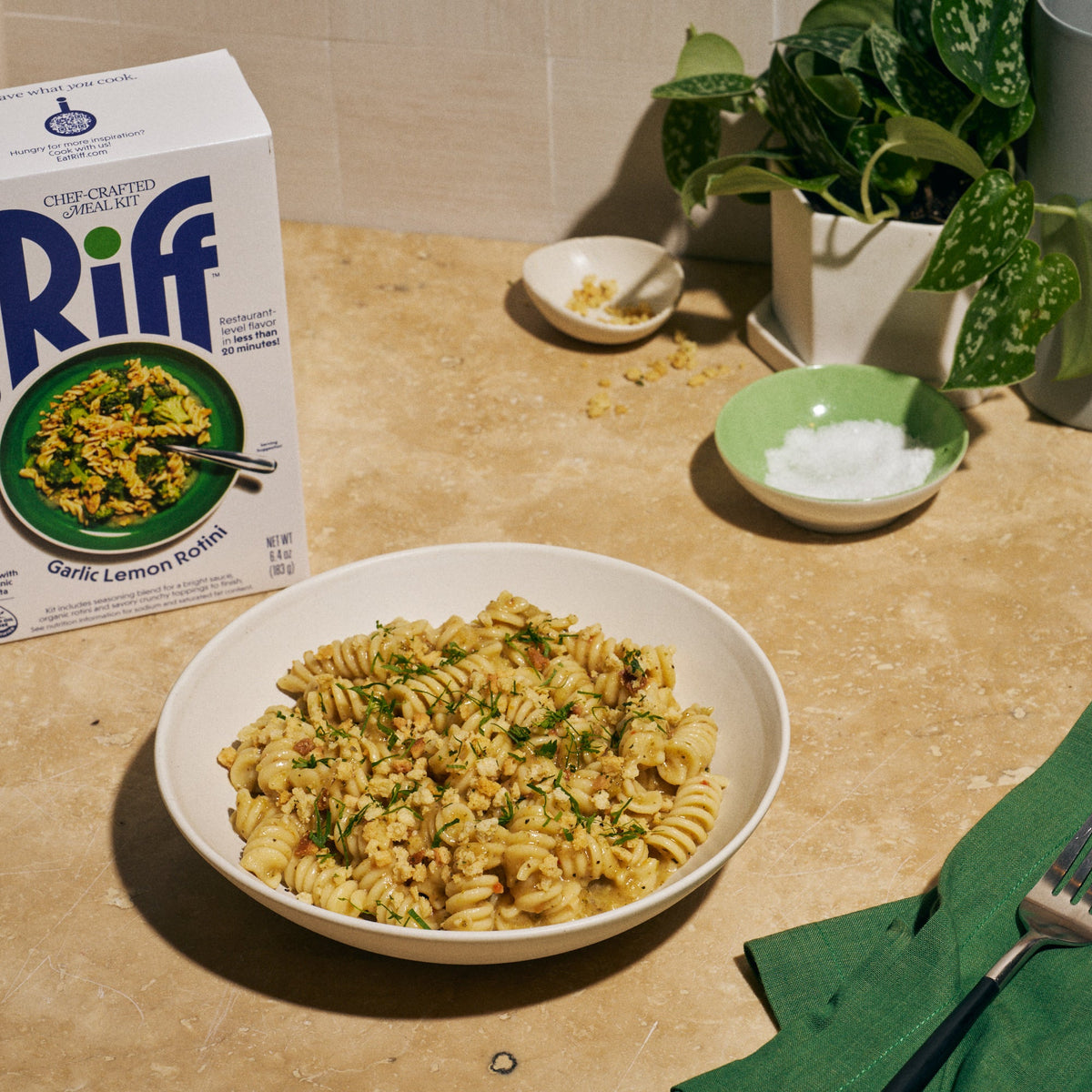 A white bowl of Riff Garlic Lemon Rotini pasta topped with crunchy breadcrumbs and fresh herbs, set on a kitchen counter next to a box of Riff Garlic Lemon Rotini chef-crafted meal kit, small dishes of salt and crunchy topping, and a potted plant.