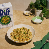 A white bowl of Riff Garlic Lemon Rotini pasta topped with crunchy breadcrumbs and fresh herbs, set on a kitchen counter next to a box of Riff Garlic Lemon Rotini chef-crafted meal kit, small dishes of salt and crunchy topping, and a potted plant.