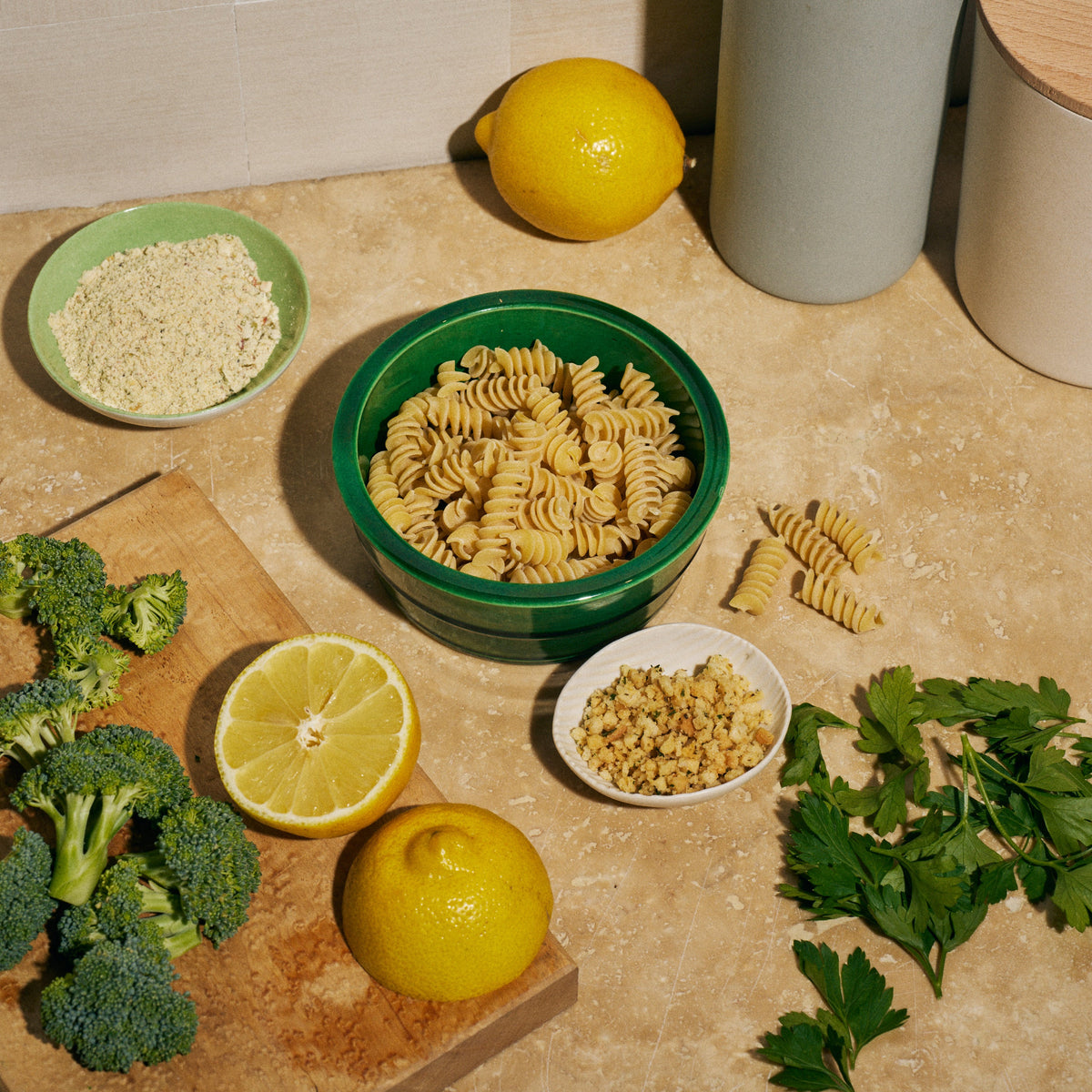 Overhead shot of ingredients on a kitchen counter for Riff Garlic Lemon Rotini, including a green bowl of raw rotini pasta, fresh broccoli florets, lemons, parsley, a bowl of seasoning, and a bowl of crunchy topping.