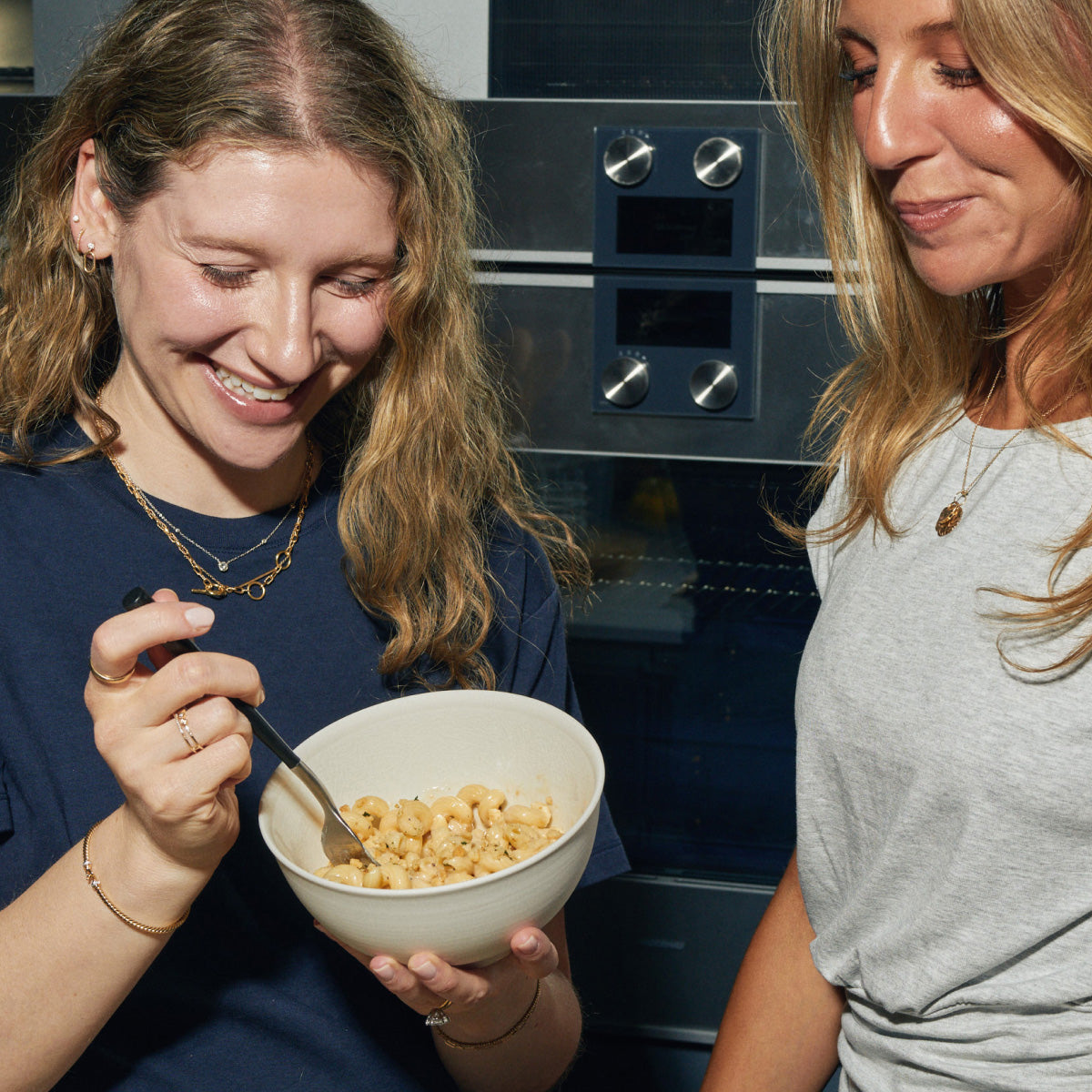 Two smiling women stand close together, one eating macaroni pasta from a white bowl with a fork while the other looks on, in a kitchen with a stainless steel oven in the background.