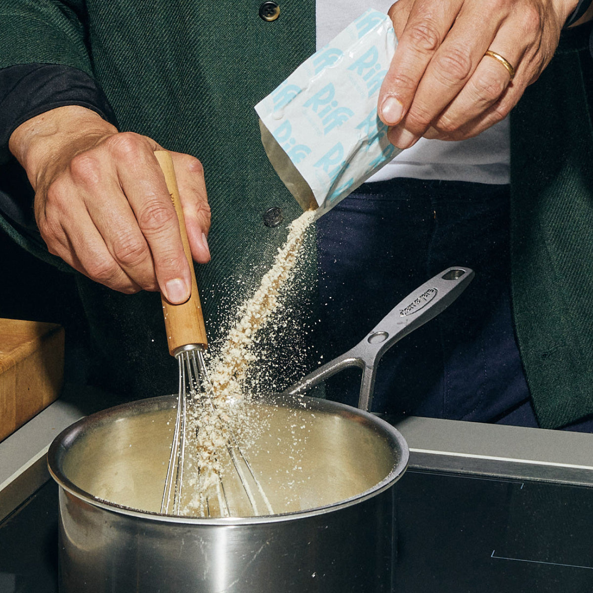 Close-up of a person's hands pouring Riff seasoning powder from a packet into a steel pot while whisking with a wooden-handled whisk.