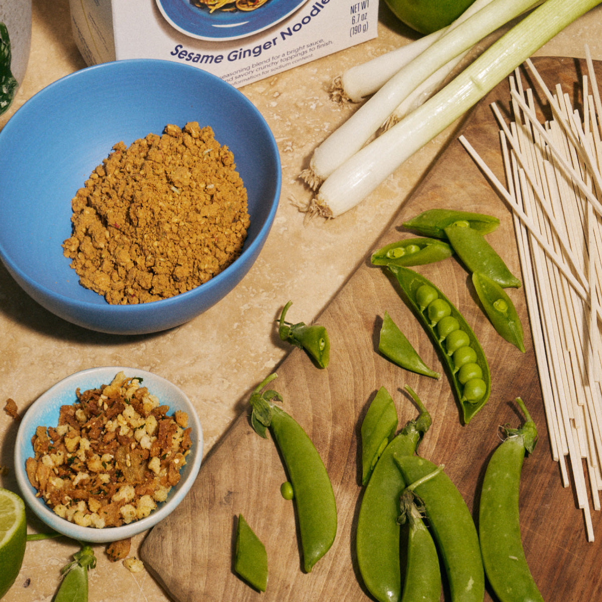 Overhead shot of a Riff Sesame Ginger Noodles meal kit box, a blue bowl of seasoning blend, a small bowl of crunchy topping, fresh scallions, snap peas, and wooden chopsticks arranged on a kitchen counter and cutting board.