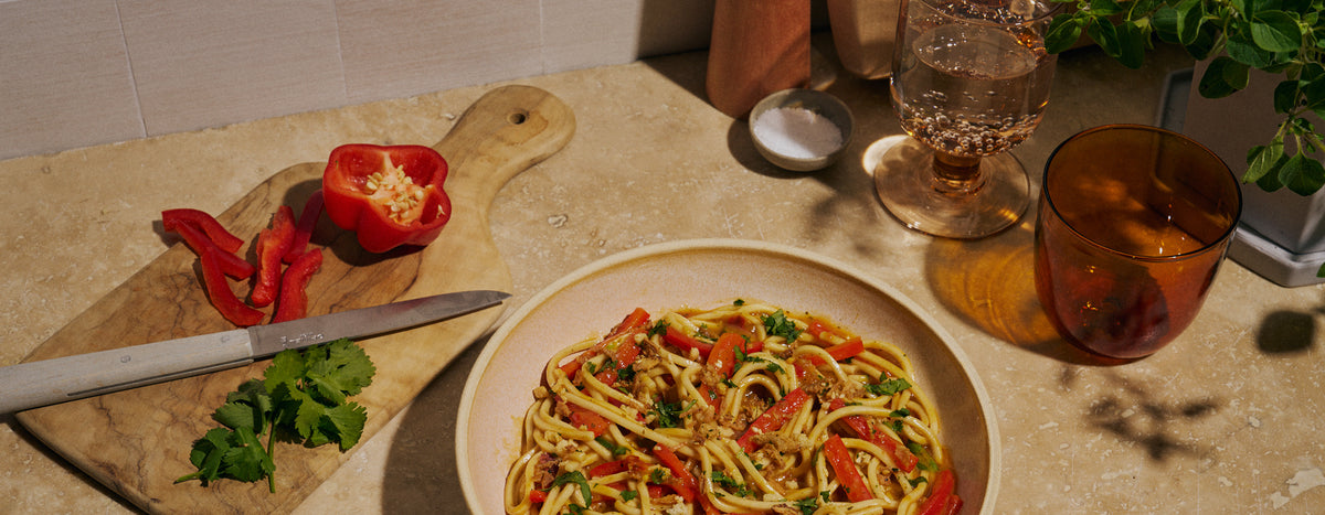 A tan bowl of noodles topped with red bell pepper strips and chopped cilantro sits on a sunlit kitchen counter next to a cutting board with a halved red bell pepper, a paring knife, a small dish of salt, and glasses of sparkling water.