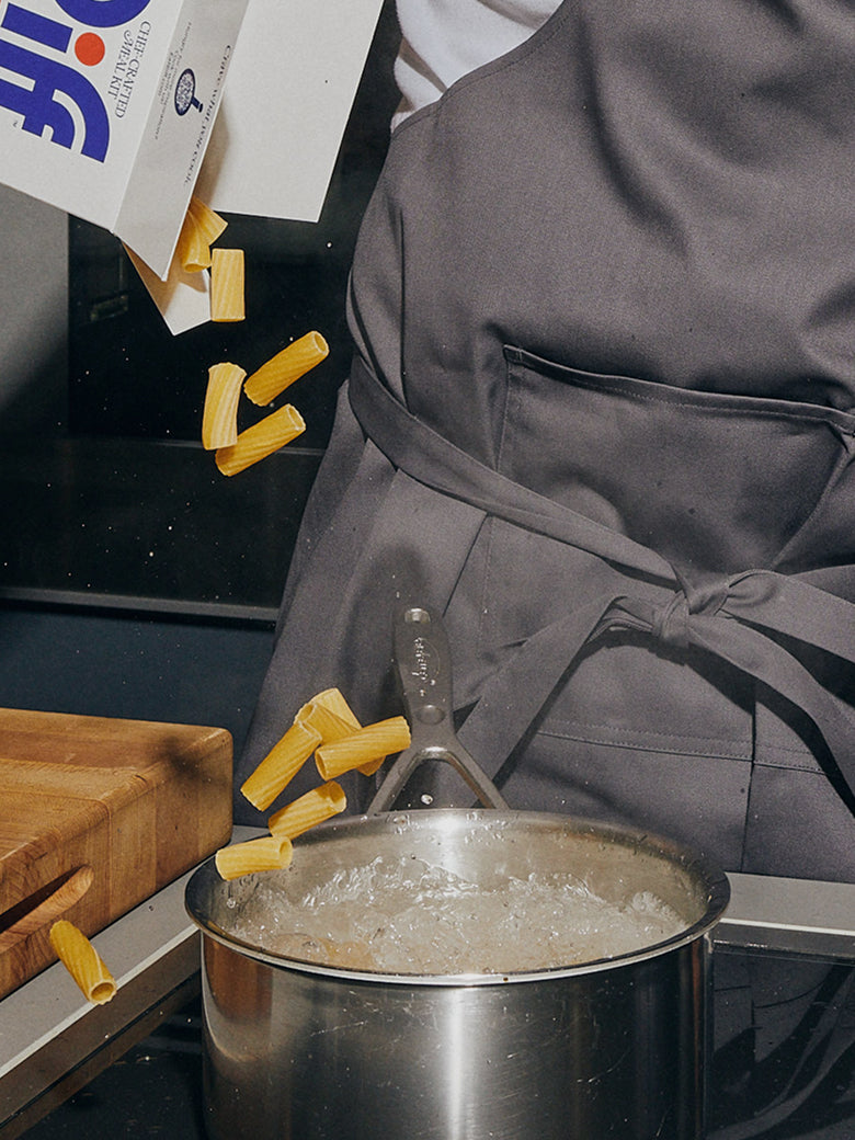 A box of Riff Spicy Tomato Rigatoni chef-crafted meal kit tips dry rigatoni pasta into a pot of boiling water, held by a person in a gray apron standing at a stovetop.