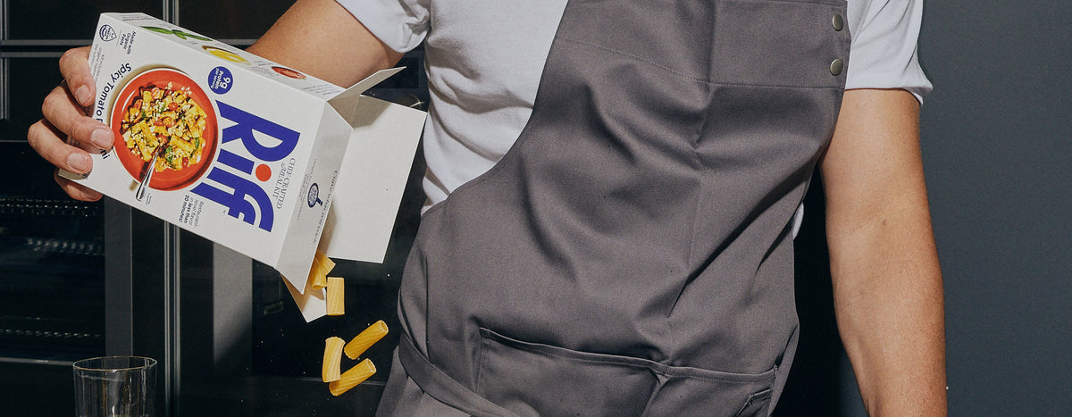 A person wearing a gray apron pours dry rigatoni pasta from an open box of Riff Spicy Tomato Rigatoni chef-crafted meal kit into a glass below.