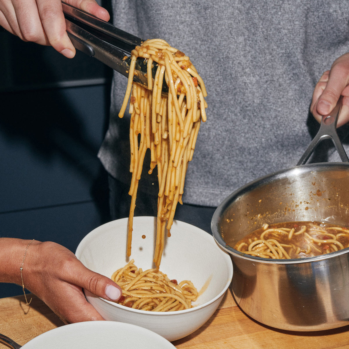 Tongs lift a portion of Riff Sesame Ginger Noodles in sauce from a metal pot into a white bowl on a wooden cutting board.