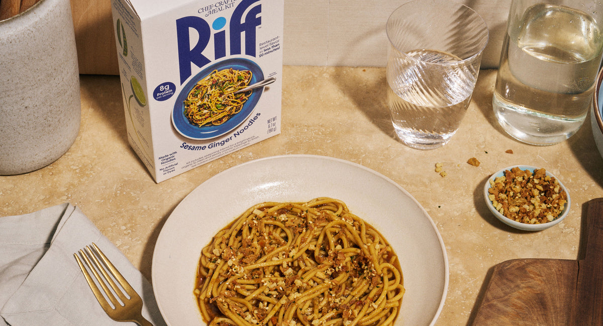 A bowl of Riff Sesame Ginger Noodles sits on a kitchen counter next to the Riff box, two glasses of water, a small dish of crunchy topping, and a gold fork on a napkin.