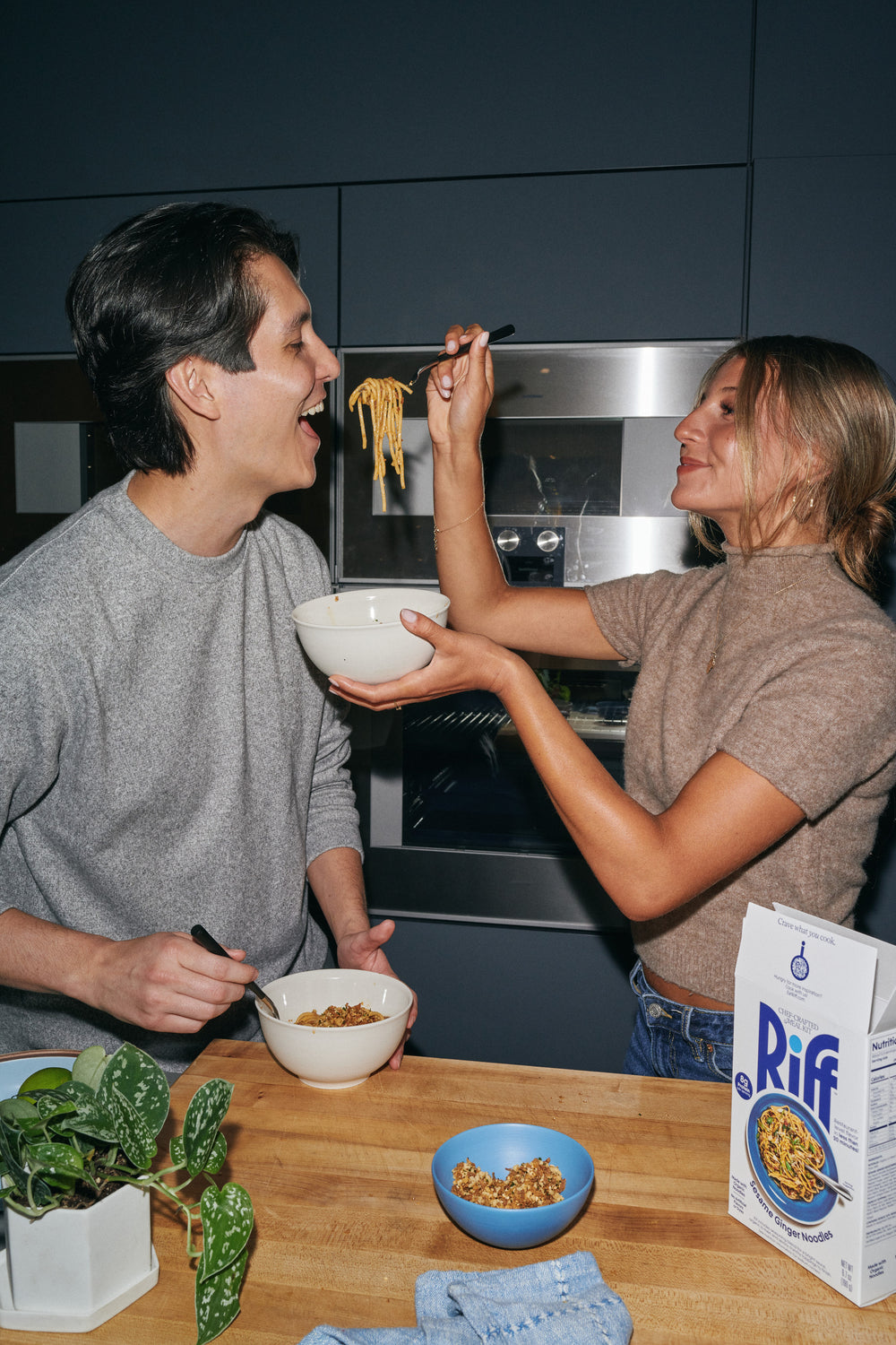 A woman feeds a forkful of Riff Sesame Ginger Noodles to a man across a kitchen counter, both smiling, with bowls of noodles and crunchy topping and a Riff box on the table.