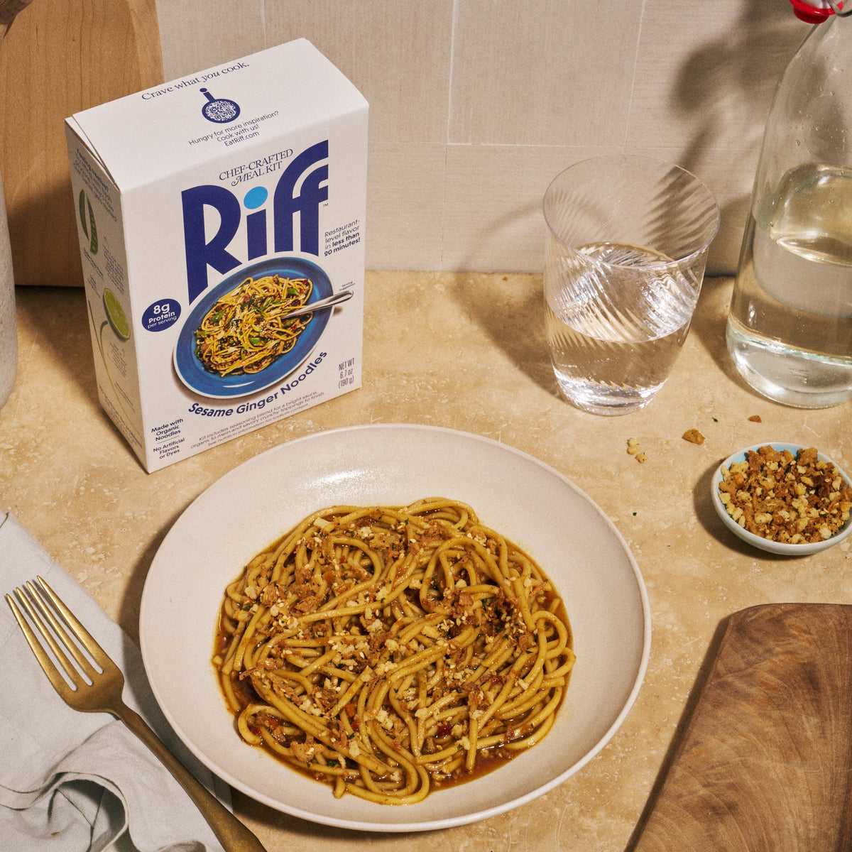 A bowl of Riff sesame ginger noodles on a kitchen counter next to the Riff box, a glass of water, a bottle of water, and a small dish of crunchy topping.