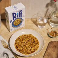 A bowl of Riff sesame ginger noodles on a kitchen counter next to the Riff box, a glass of water, a bottle of water, and a small dish of crunchy topping.