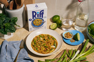 A white bowl of Riff Sesame Ginger Noodles topped with scallions and crispy shallots sits on a kitchen counter next to a box of Riff Sesame Ginger Noodles chef-crafted meal kit, fresh limes, a glass of water, a bottle of sparkling water, snap peas, and scallions.
