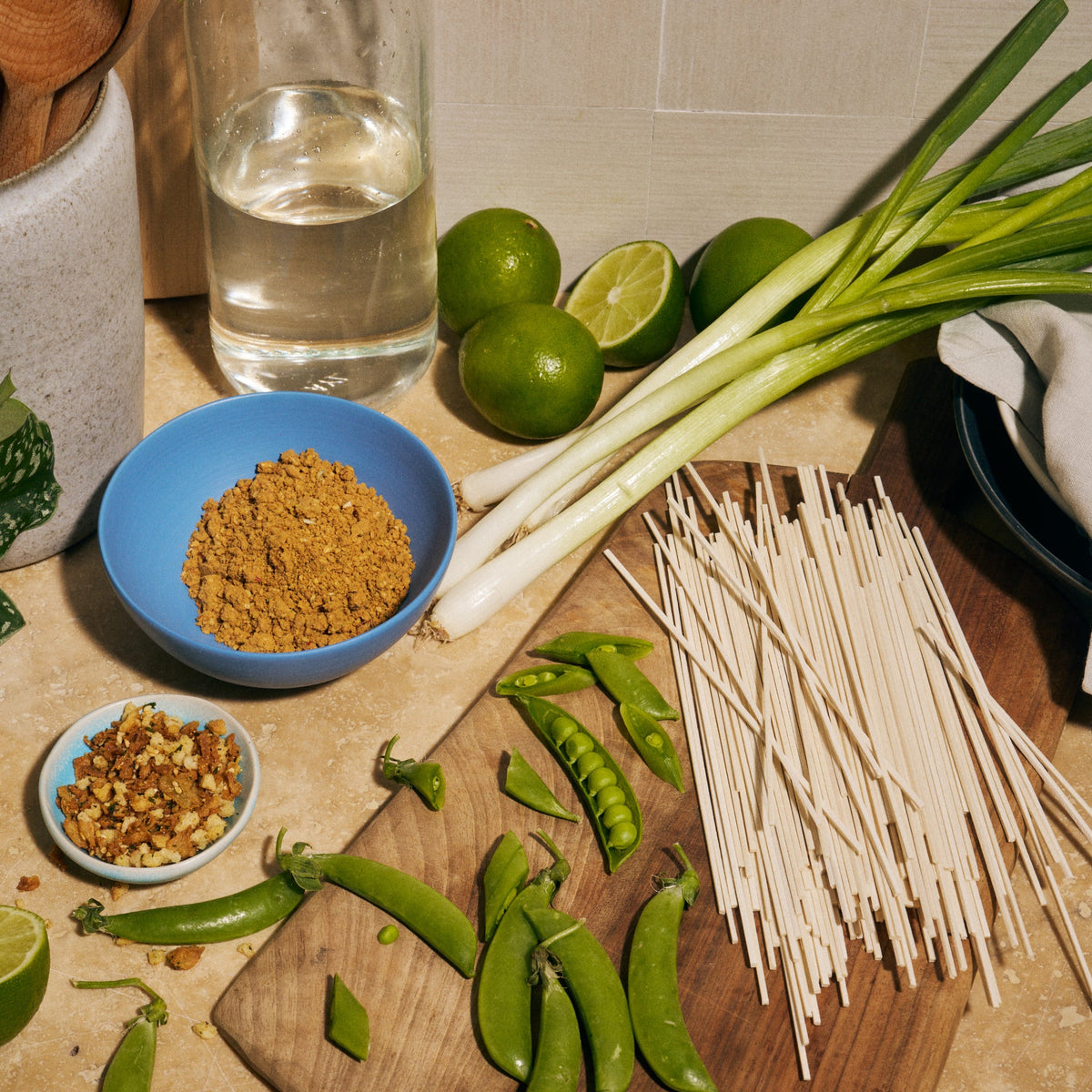 Overhead shot of ingredients on a kitchen counter for Riff Sesame Ginger Noodles, including dry lo mein noodles, scallions, limes, snap peas, a bowl of seasoning, and a bowl of crunchy topping.