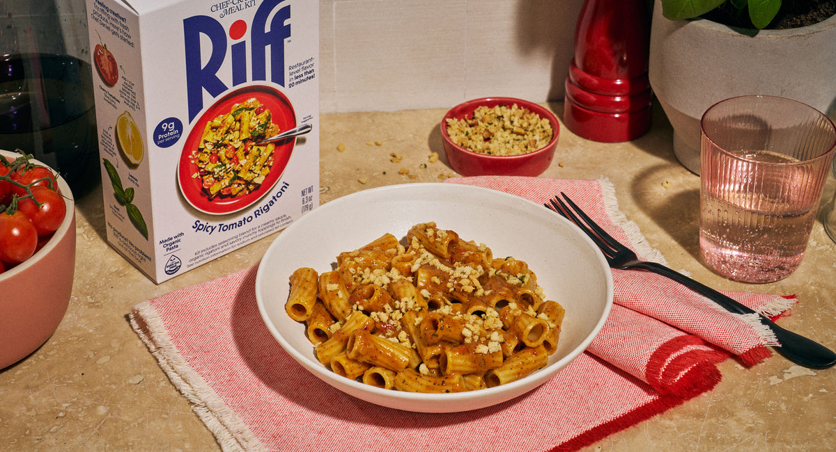 A white bowl of Riff Spicy Tomato Rigatoni topped with crunchy breadcrumbs sits on a red napkin next to a black fork, with the Riff box, a bowl of cherry tomatoes, a bowl of crunchy topping, a red pepper grinder, and a glass of pink beverage on the counter.