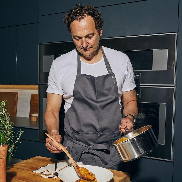 A curly-haired man wearing a white t-shirt and gray apron plates pasta from a steel pot onto a white dish with a wooden spoon in a dark kitchen.