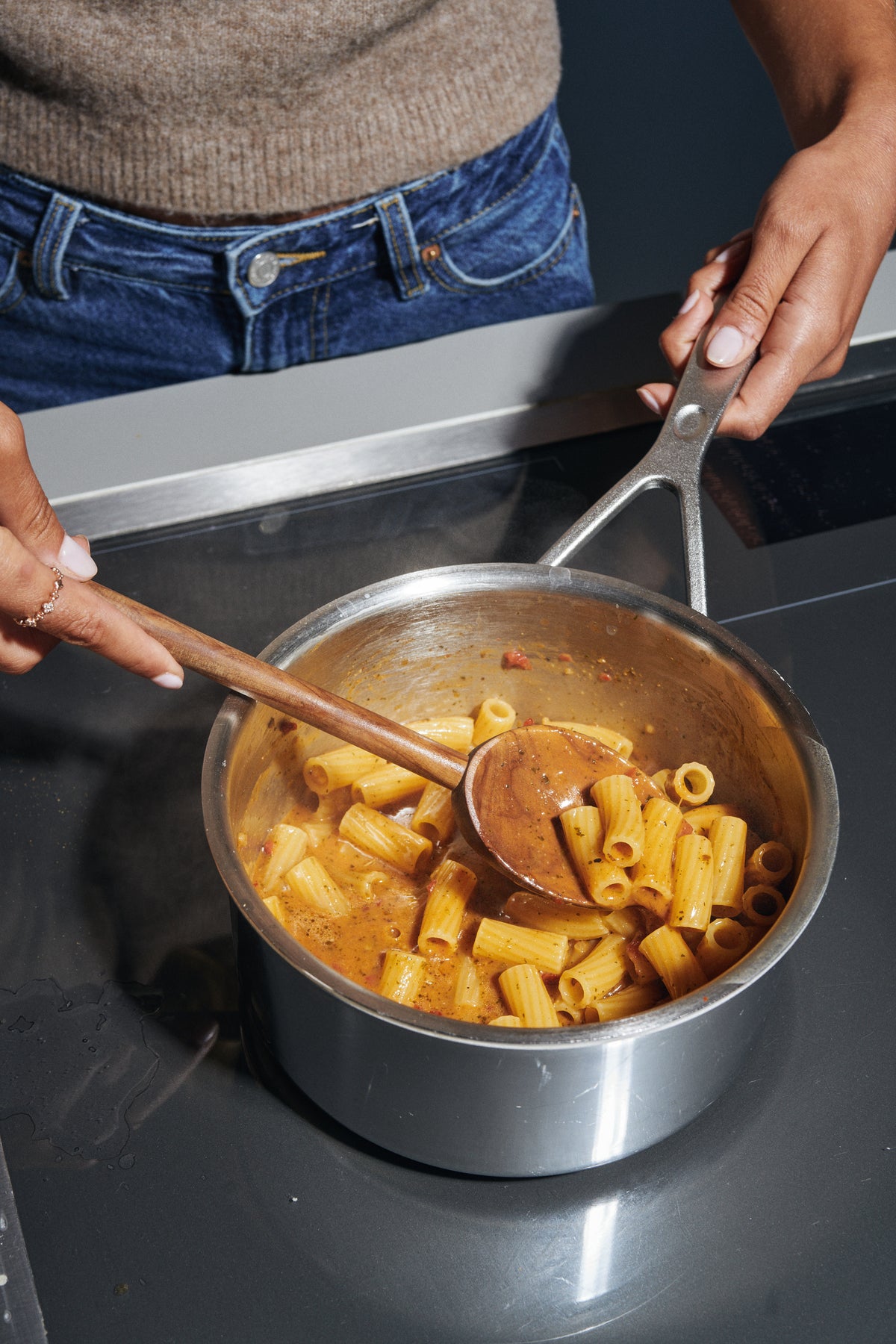 A stainless steel saucepan filled with Riff Rigatoni pasta in a creamy orange sauce on a dark stovetop, with a person's hands holding the handles and a wooden spoon stirring the pasta.