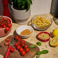 Overhead shot of ingredients on a kitchen counter for Riff Spicy Tomato Rigatoni, including a glass bowl of raw rigatoni pasta, cherry tomatoes on the vine, a lemon, basil, a pepper grinder, a bowl of seasoning, and a bowl of crunchy topping.