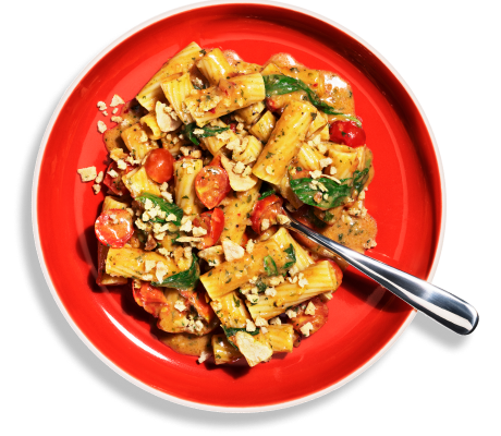Overhead shot of Riff Spicy Tomato Rigatoni topped with cherry tomatoes, basil, and a crunchy topping in a red bowl, with a fork resting on the side.
