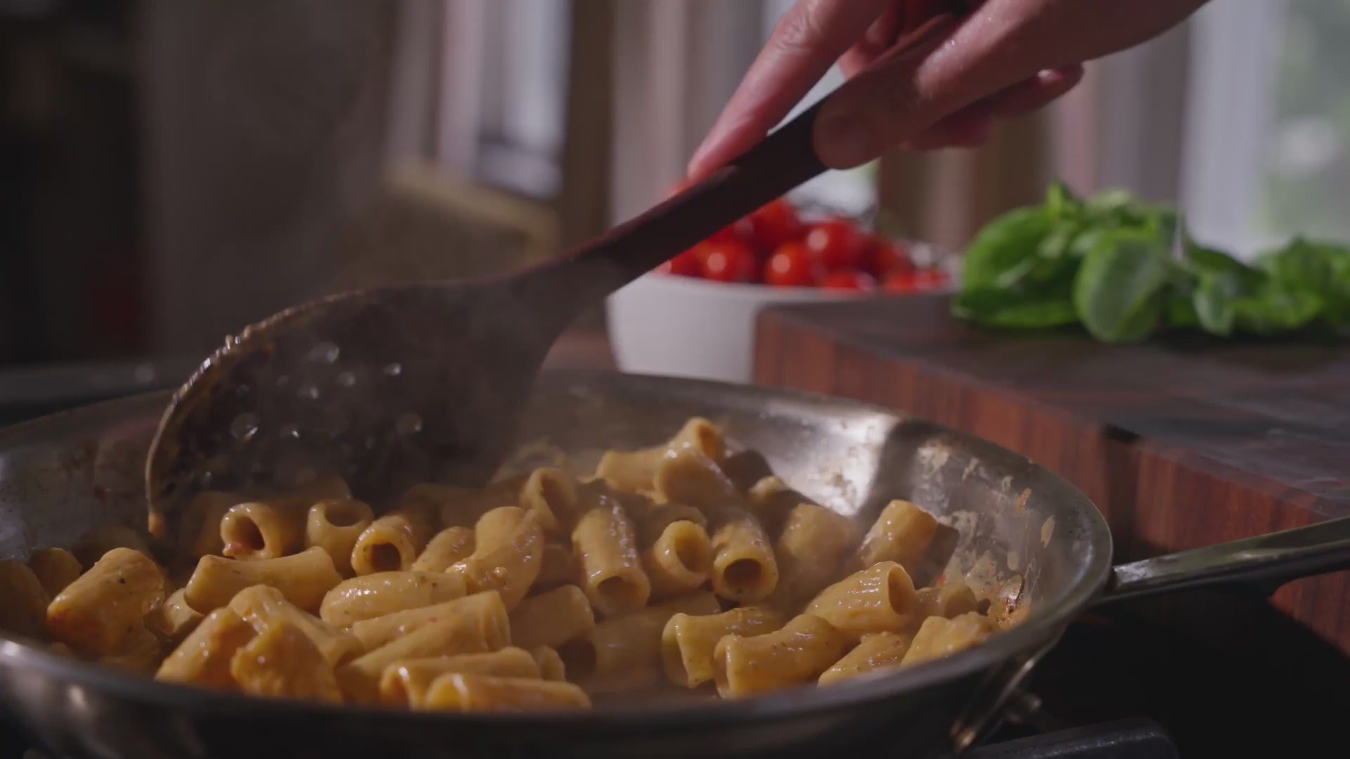 Video of a wooden spoon stirring Riff rigatoni pasta in a steaming skillet, with cherry tomatoes and basil on a cutting board in the background.