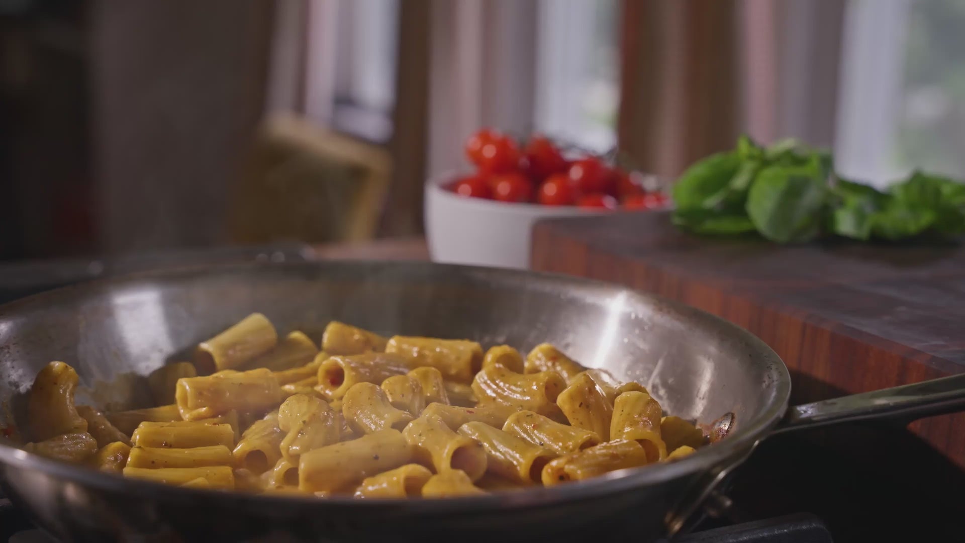 Video of steam rising from a skillet of Riff rigatoni pasta in sauce, with a bowl of cherry tomatoes and fresh basil on the counter behind it.