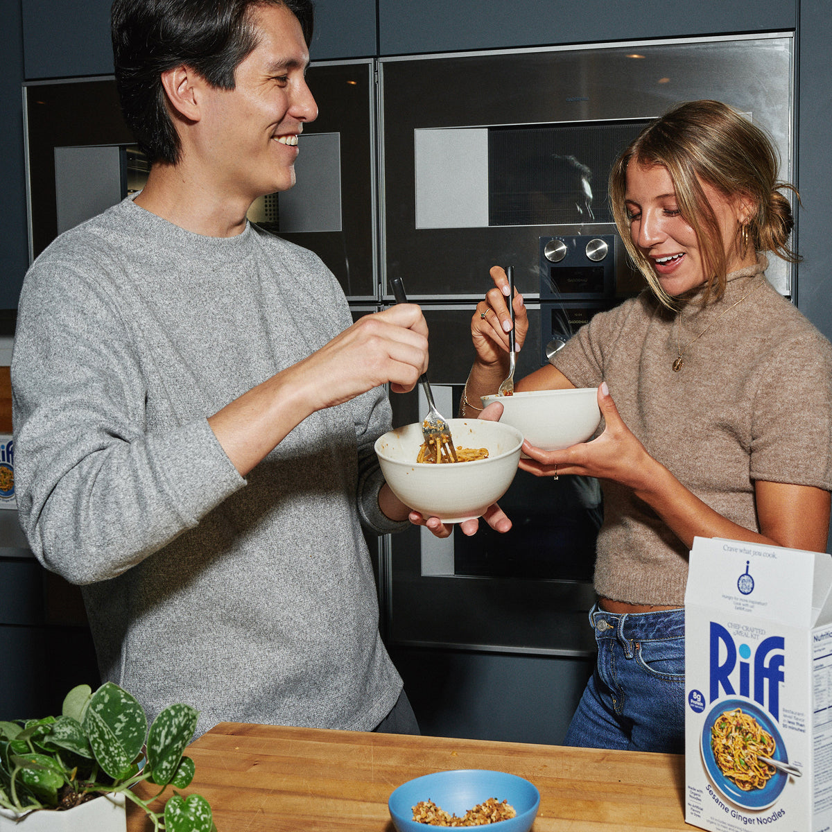 A man and woman in a kitchen each hold a white bowl of noodles and eat with forks, smiling at each other, with a box of Riff Sesame Ginger Noodles chef-crafted meal kit and a blue bowl of crunchy topping on the counter.